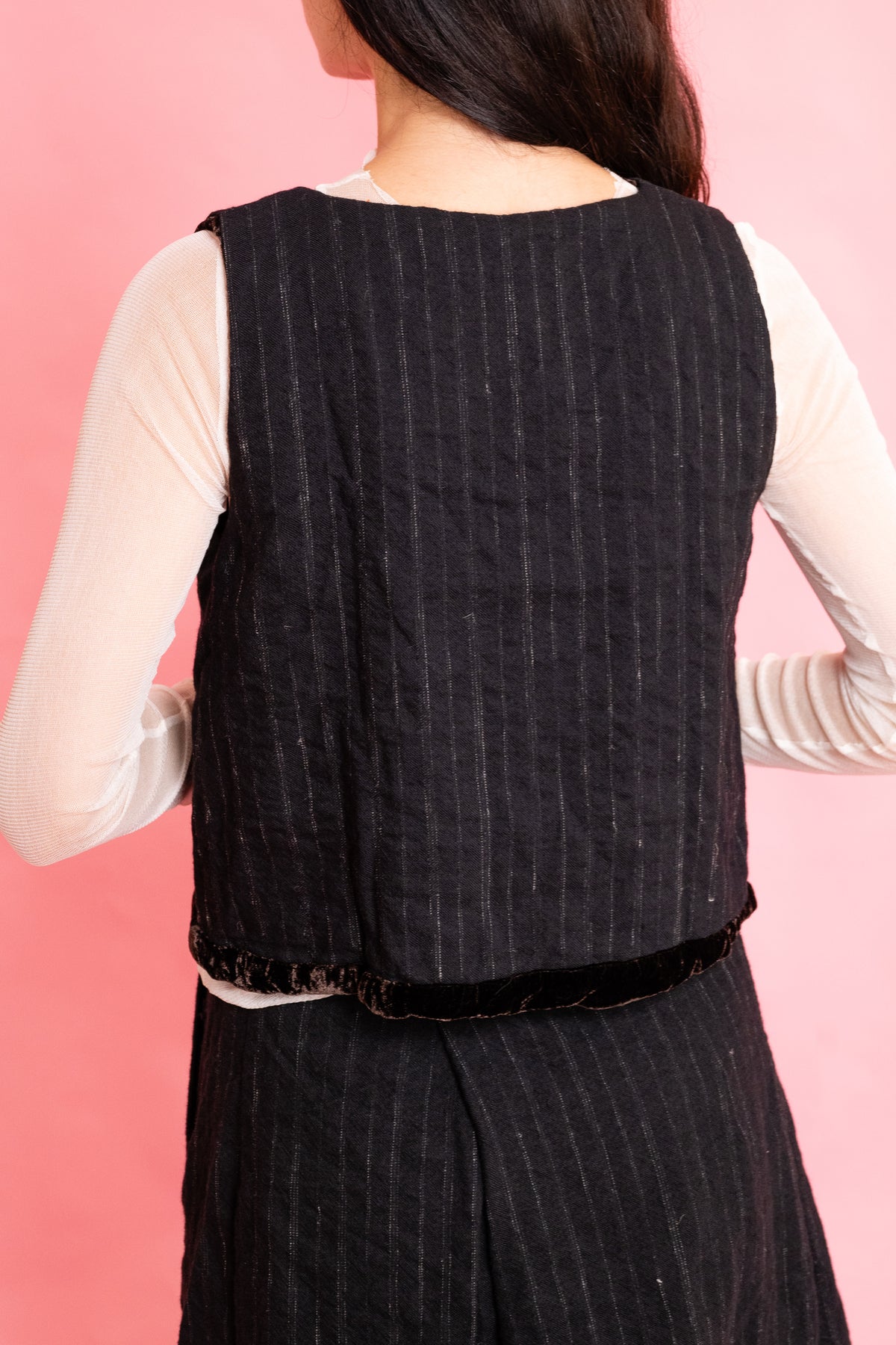 Back view of model wearing a wool stripe buttoned waistcoat with brown velvet trim with a cream mesh long sleeve tee shirt and panelled wool stripe skirt on a pink background