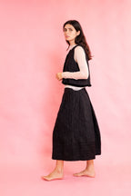 Side view of model wearing a wool stripe buttoned waistcoat with brown velvet trim with a cream mesh long sleeve tee shirt and panelled wool stripe skirt on a pink background