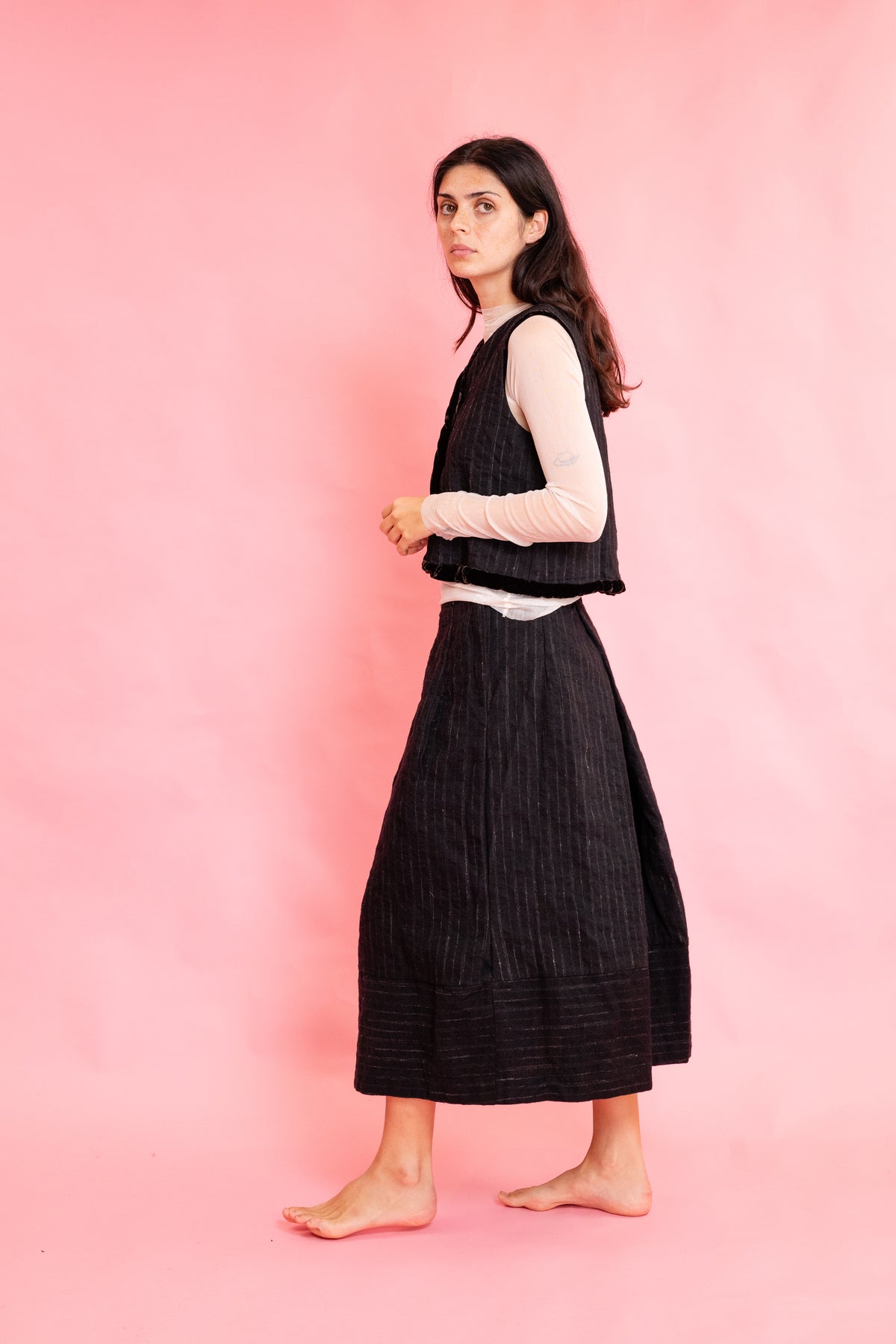 Side view of model wearing a wool stripe buttoned waistcoat with brown velvet trim with a cream mesh long sleeve tee shirt and panelled wool stripe skirt on a pink background
