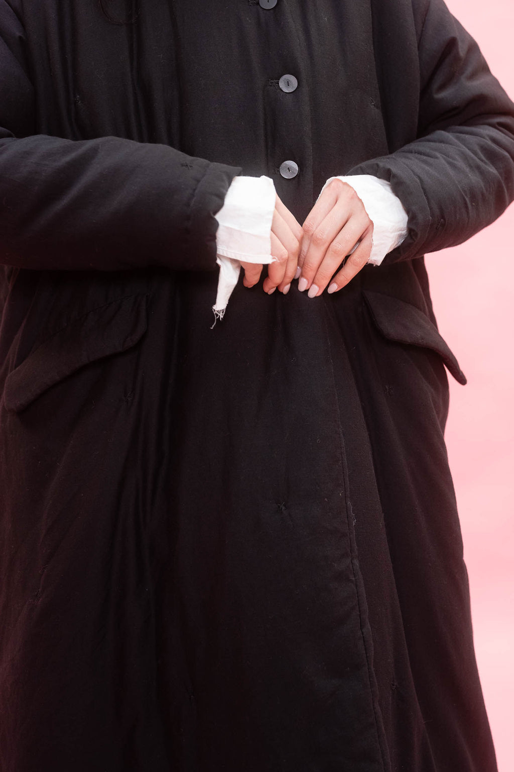 A close-up of a woman wearing a long black coat with white cuffs, standing against a solid pink background.