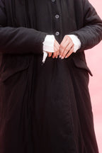 A close-up of a woman wearing a long black coat with white cuffs, standing against a solid pink background.