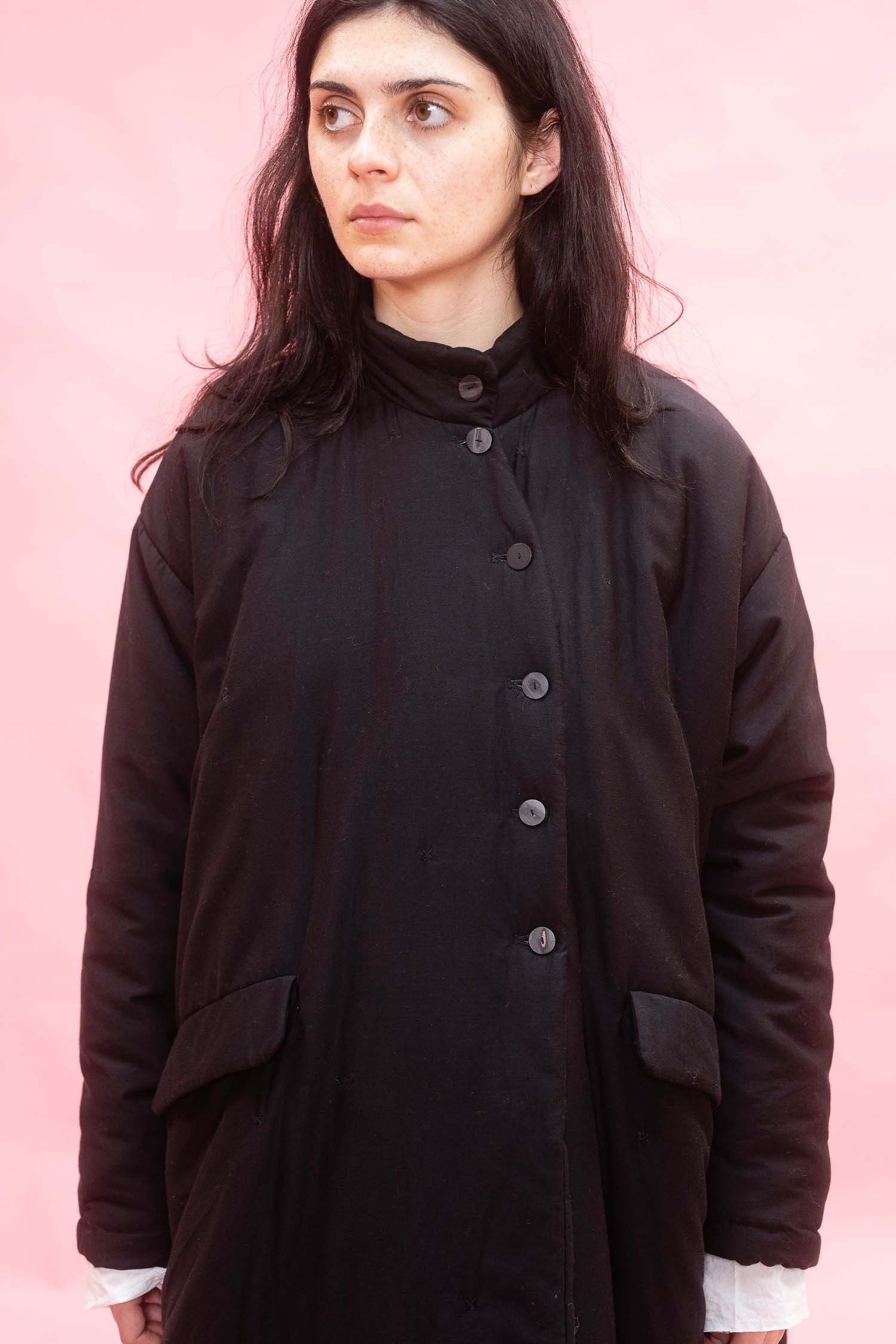 A woman with long dark hair wearing a black button-front coat over a white shirt, standing against a solid pink background.