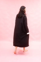 A woman with long dark hair is shown from behind wearing a long black quilted coat with a white cuff detail, standing barefoot against a solid pink backdrop.