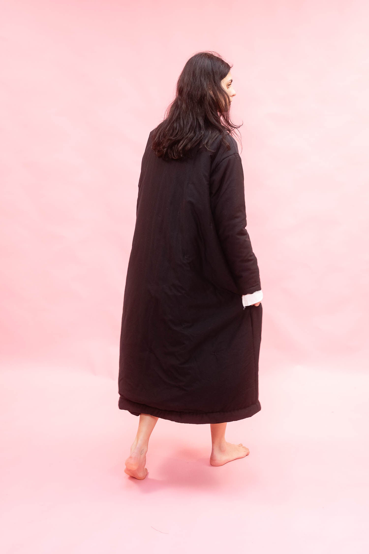 A woman with long dark hair is shown from behind wearing a long black quilted coat with a white cuff detail, standing barefoot against a solid pink backdrop.