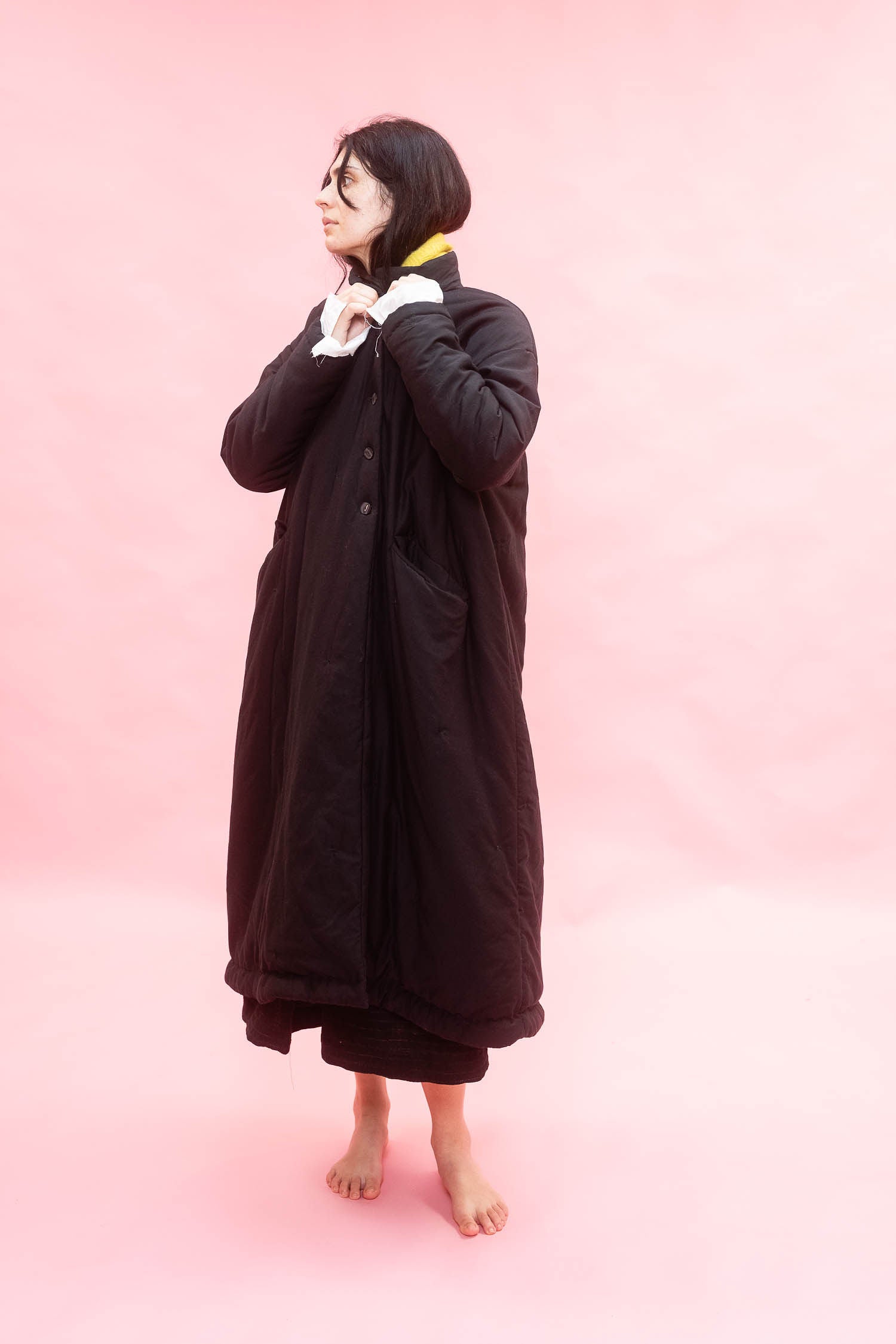 A woman stands barefoot against a solid pink studio background, wearing a long black quilted cuddle coat with white gloves and a yellow scarf