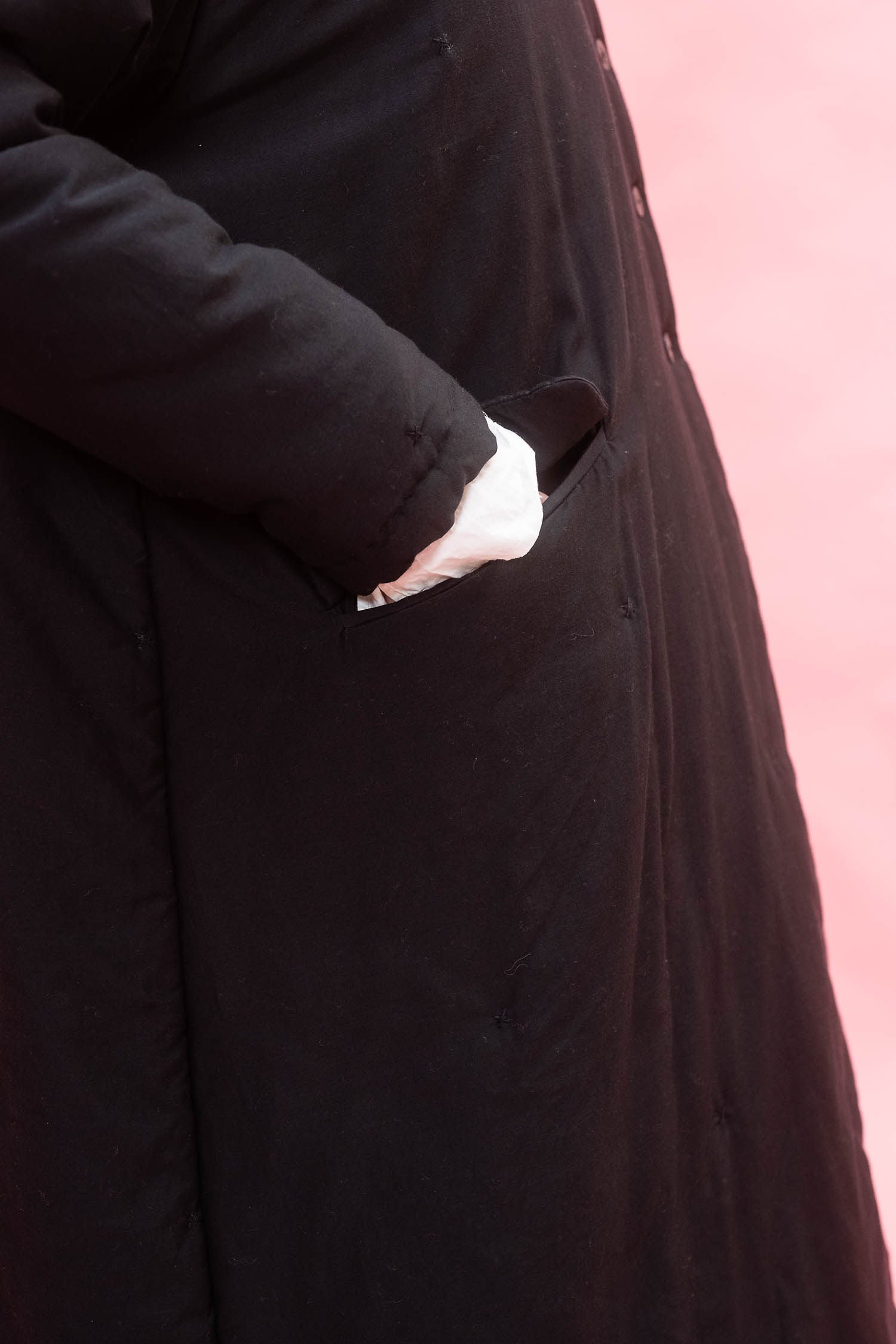 Close-up of a black quilted long-line coat with a side pocket, showing a hand reaching into the pocket, against a pink background.
