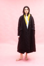 A model stands barefoot against a solid pink studio background, wearing a long black quilted cuddle over coat over a white shirt and a bright yellow scarf.