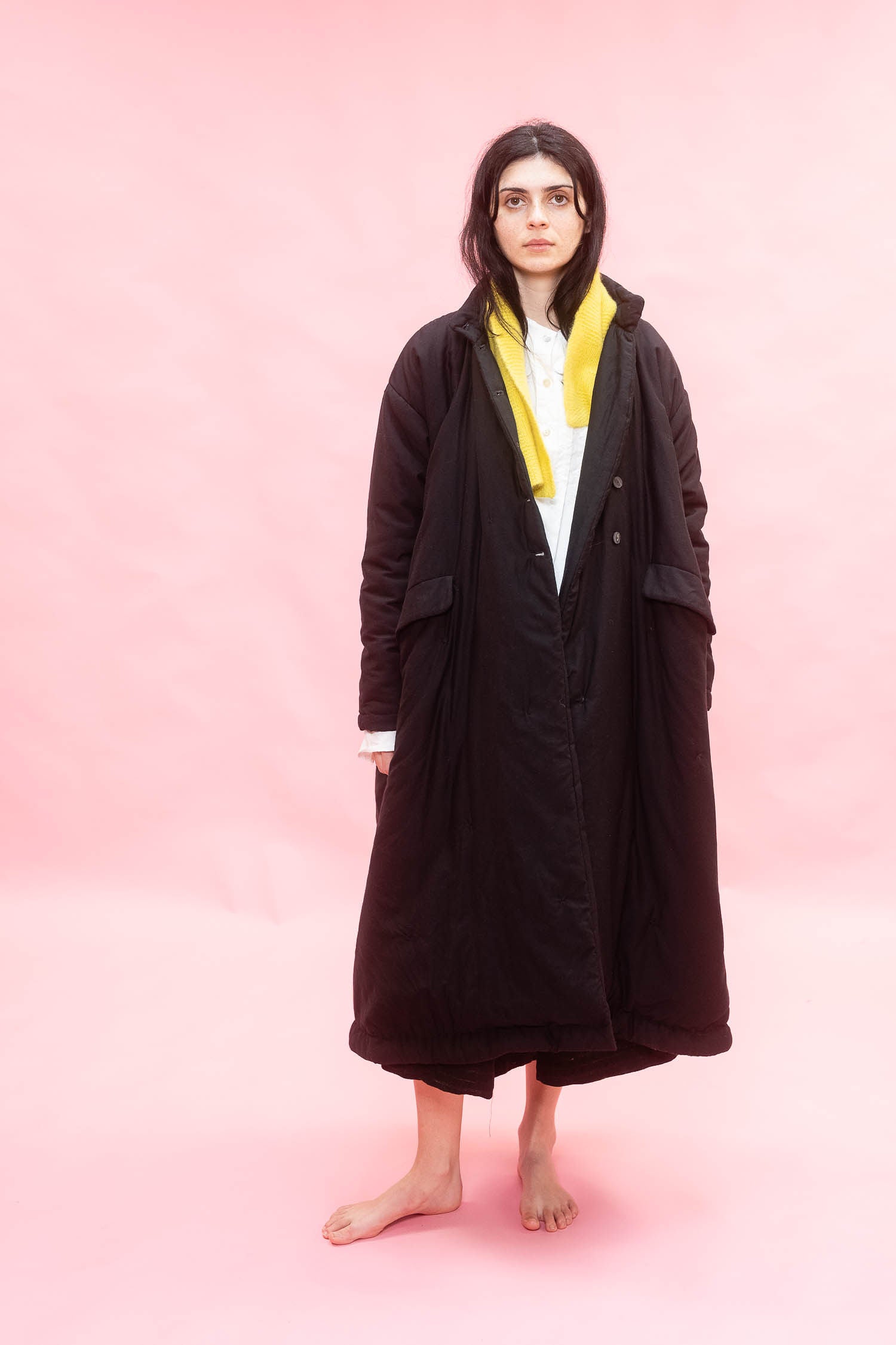 A model stands barefoot against a solid pink studio background, wearing a long black quilted cuddle over coat over a white shirt and a bright yellow scarf.