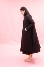 A woman stands barefoot in profile wearing a long black quilted coat against a solid pink studio background.