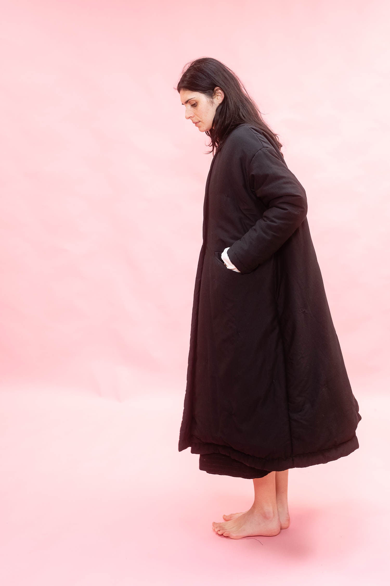 A woman stands barefoot in profile wearing a long black quilted coat against a solid pink studio background.
