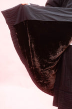 A close-up of a woman holding up a black cuddle coat showing the brown velvet contrast lining against a pale pink background.