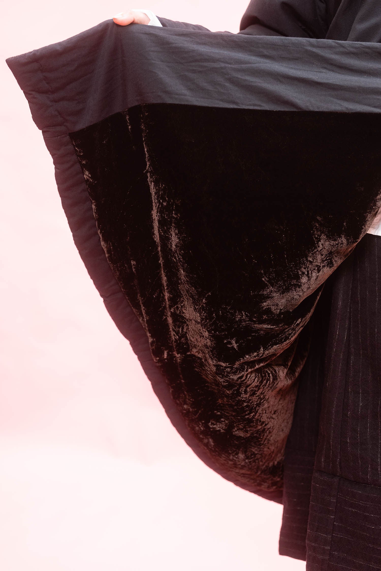 A close-up of a woman holding up a black cuddle coat showing the brown velvet contrast lining against a pale pink background.