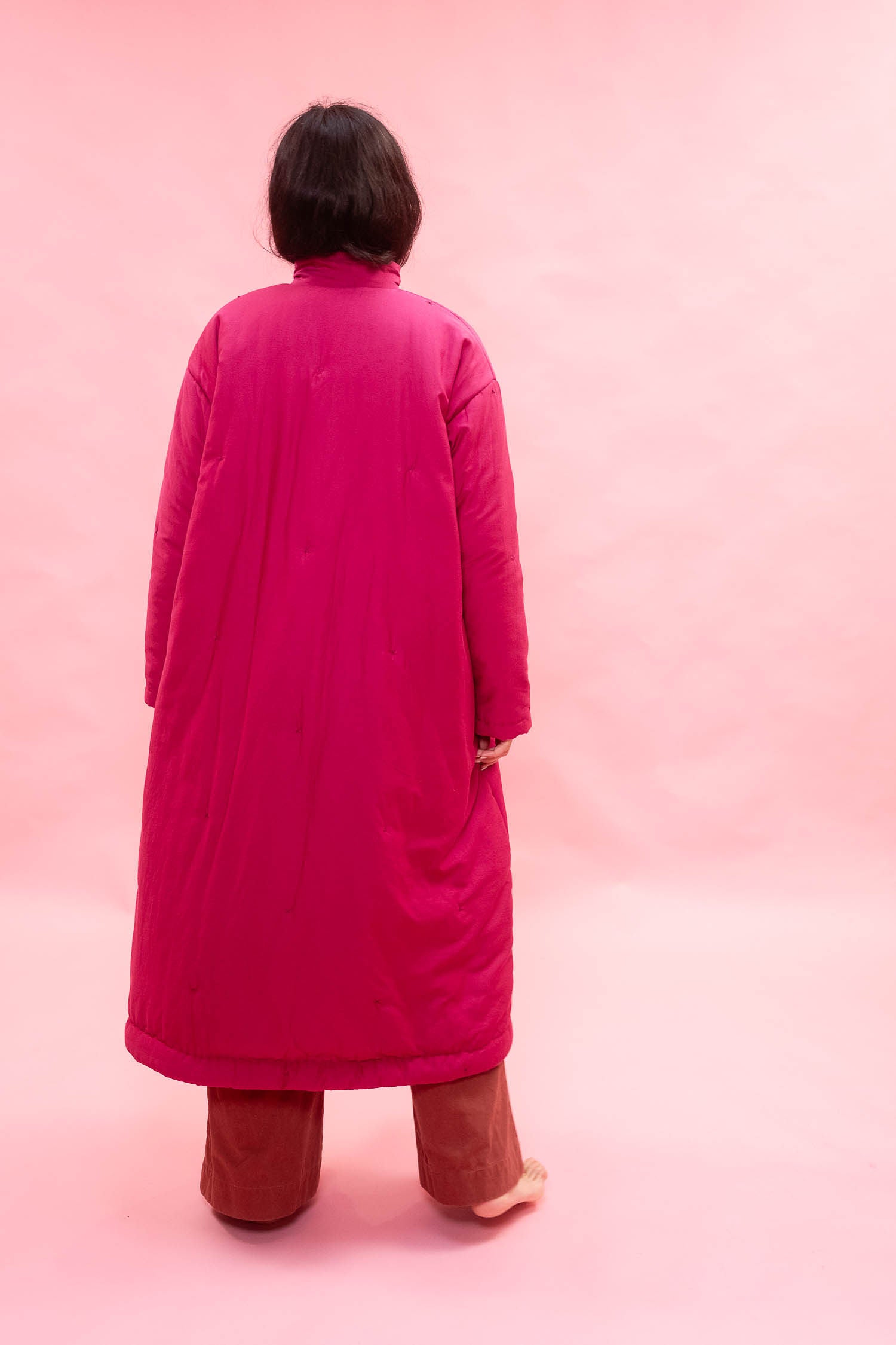 A woman viewed from behind wearing a long bright pink quilted coat over brown pants, standing against a solid pink studio background.