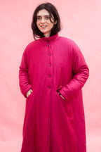 A woman wearing a bright magenta pink quilted coat with a high collar and asymmetric  front buttons, standing against a solid pink background.