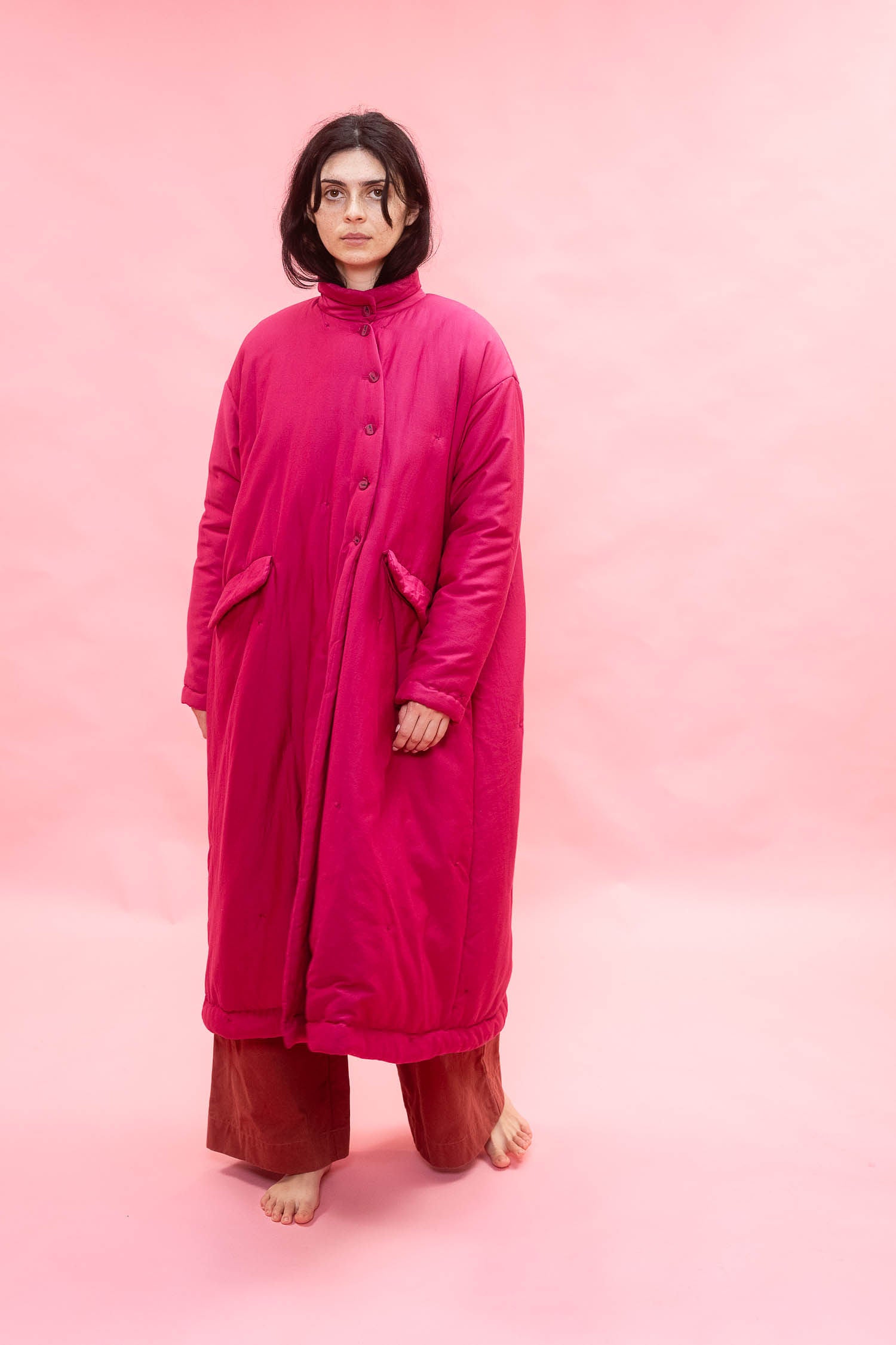 A model stands barefoot against a solid pink studio background, wearing a long, vibrant granita magenta quilted coat with a high collar and asymmetric front buttons, paired with rust-brown pants.