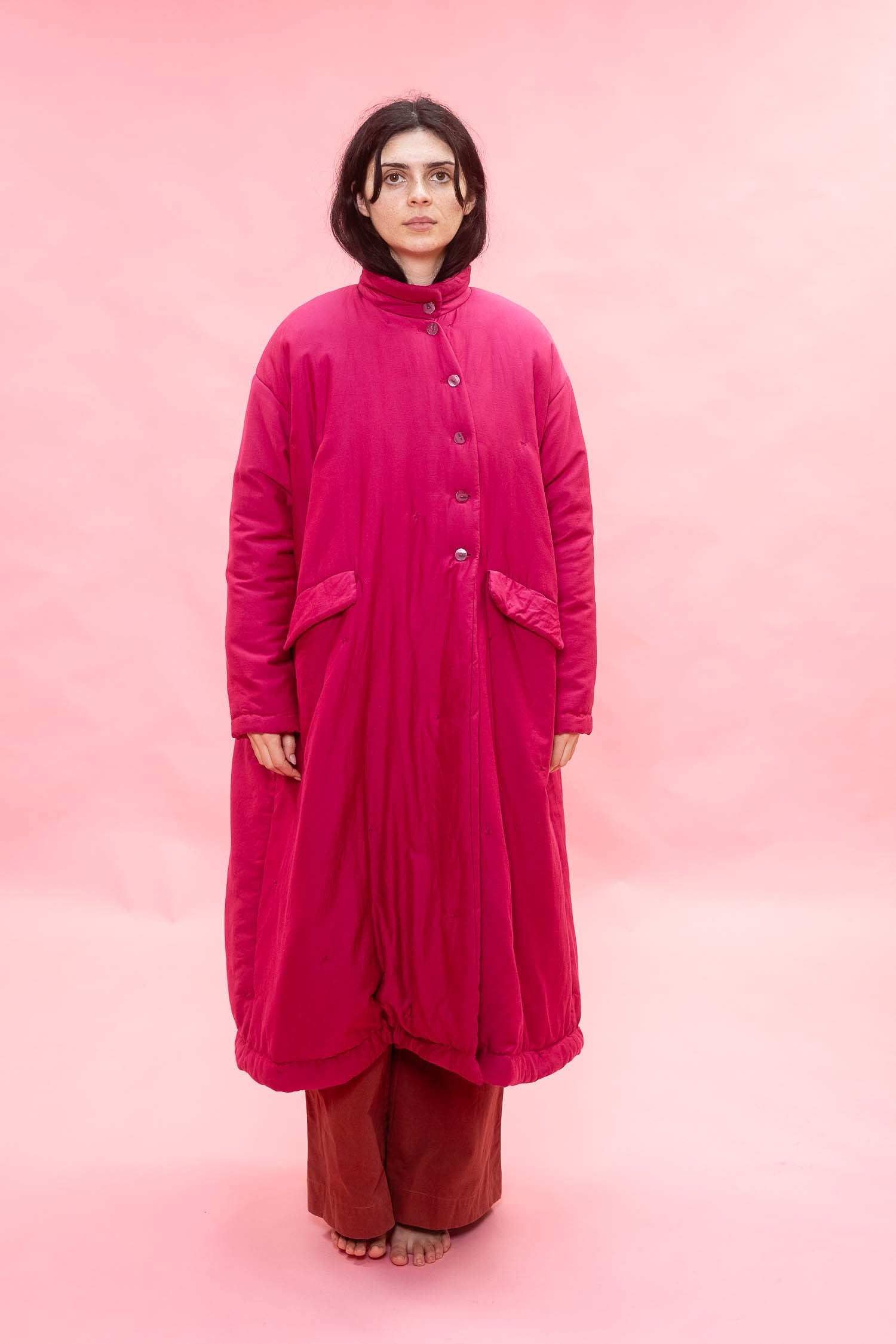 A model stands against a solid pink studio backdrop wearing a long, high-neck, button-front quilted coat in a vibrant magenta pink, paired with rust-colored pants.