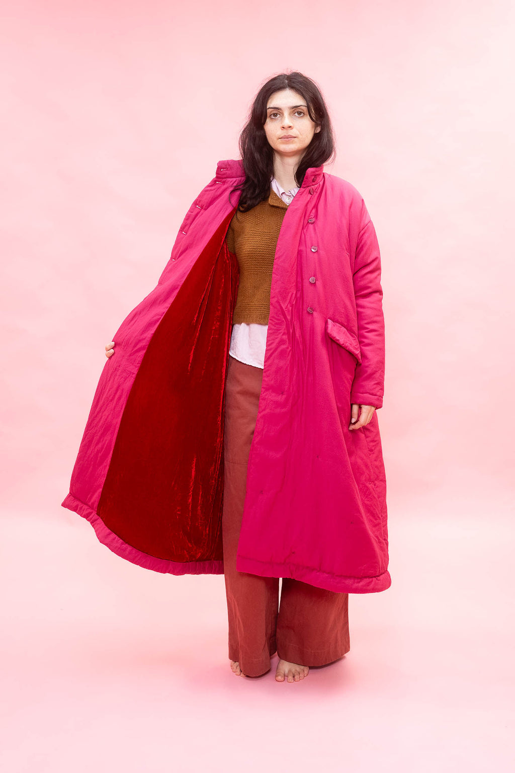A model stands barefoot against a solid pink studio background, wearing a vibrant magenta quilted coat with a contrasting deep red velvet lining. She is styled with a brown knit cardigan, white shirt, and rust-colored wide-leg pants.