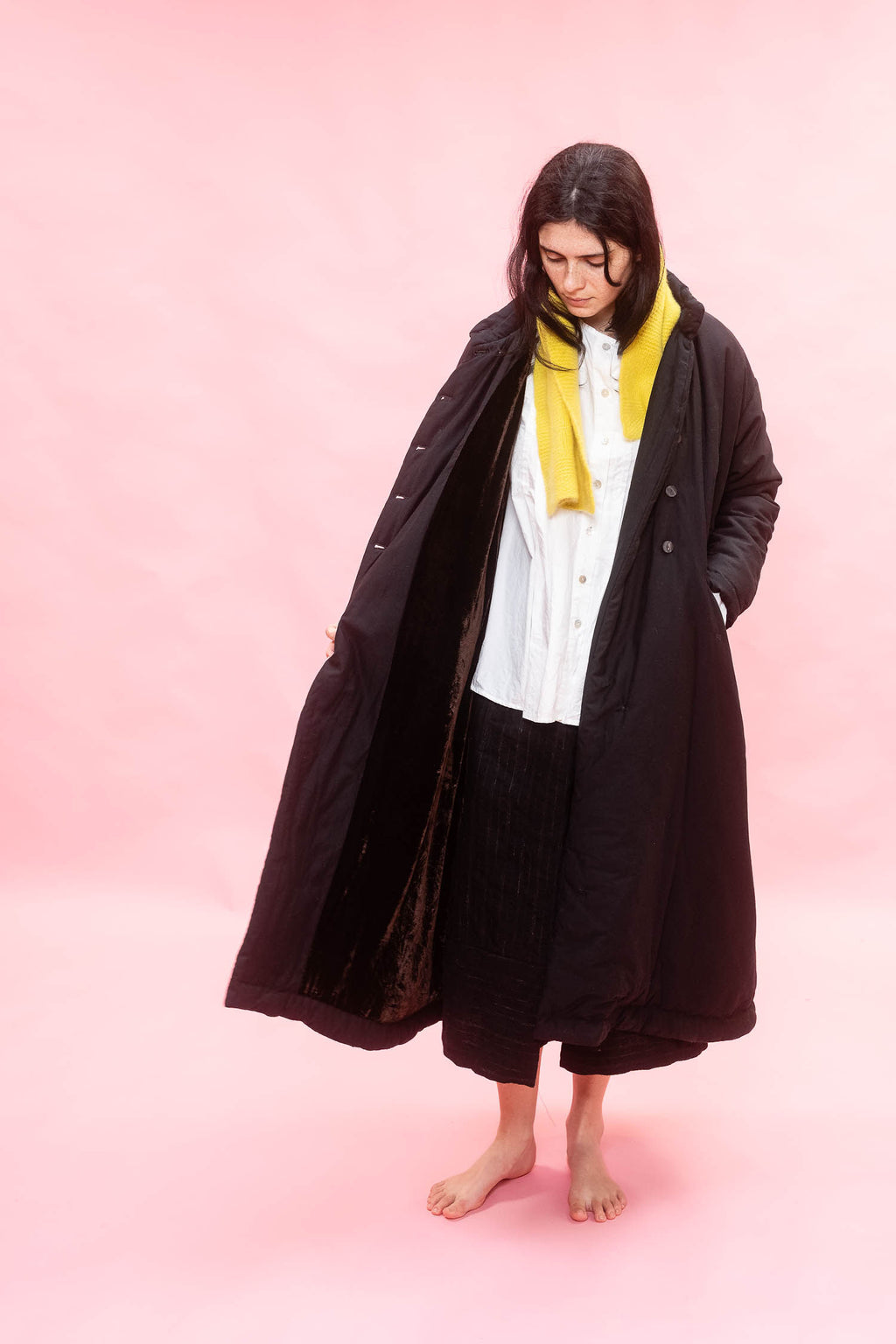 A woman stands barefoot against a solid pink studio background, wearing a long black quilted coat over a white shirt and black pleated skirt, with a bright yellow scarf wrapped around her neck.