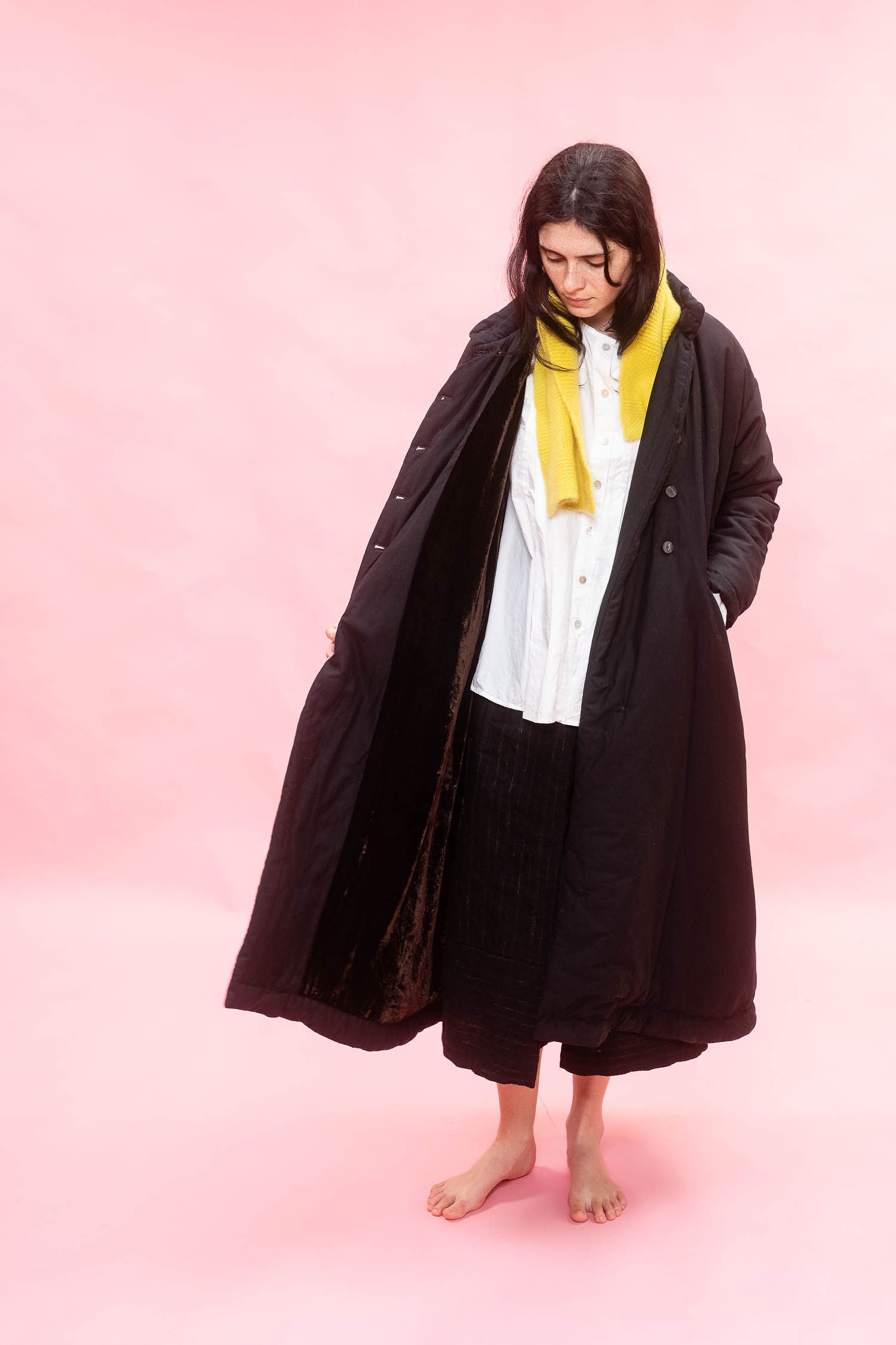 A woman stands barefoot against a solid pink studio background, wearing a long black quilted coat over a white shirt and black pleated skirt, with a bright yellow scarf wrapped around her neck.