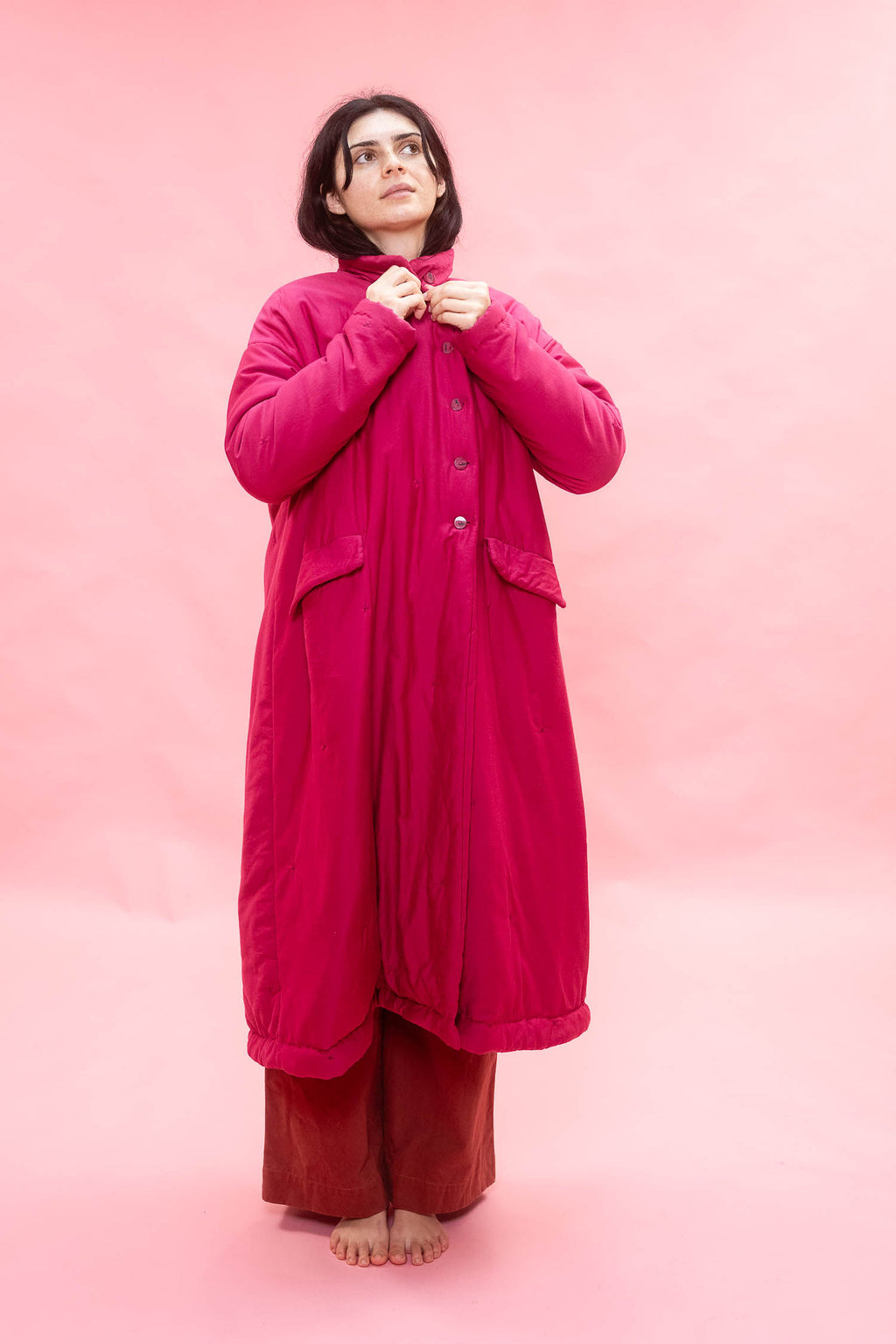 A woman stands barefoot against a solid pink studio backdrop, wearing a long bright pink quilted coat over rust-brown pants and adjusting the collar of the coat.