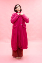 A woman stands barefoot against a solid pink studio backdrop, wearing a long bright pink quilted coat over rust-brown pants and adjusting the collar of the coat.
