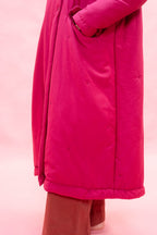 A close-up of a woman wearing a bright fuchsia pink quilted coat with side pockets, paired with brown corduroy trousers, against a solid pink background.