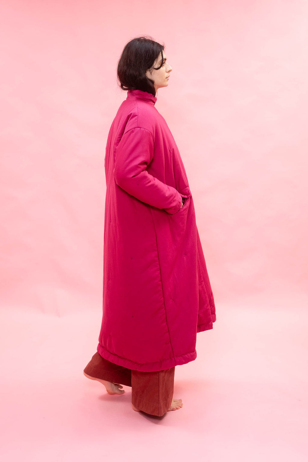 A woman wearing a long magenta-pink quilted coat over rust-brown pants, standing in profile against a solid pink studio backdrop.