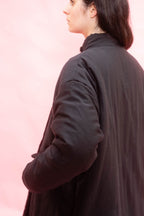 A woman shown from the back and side wearing an anthracite or black cuddle coat against a soft pink studio background.