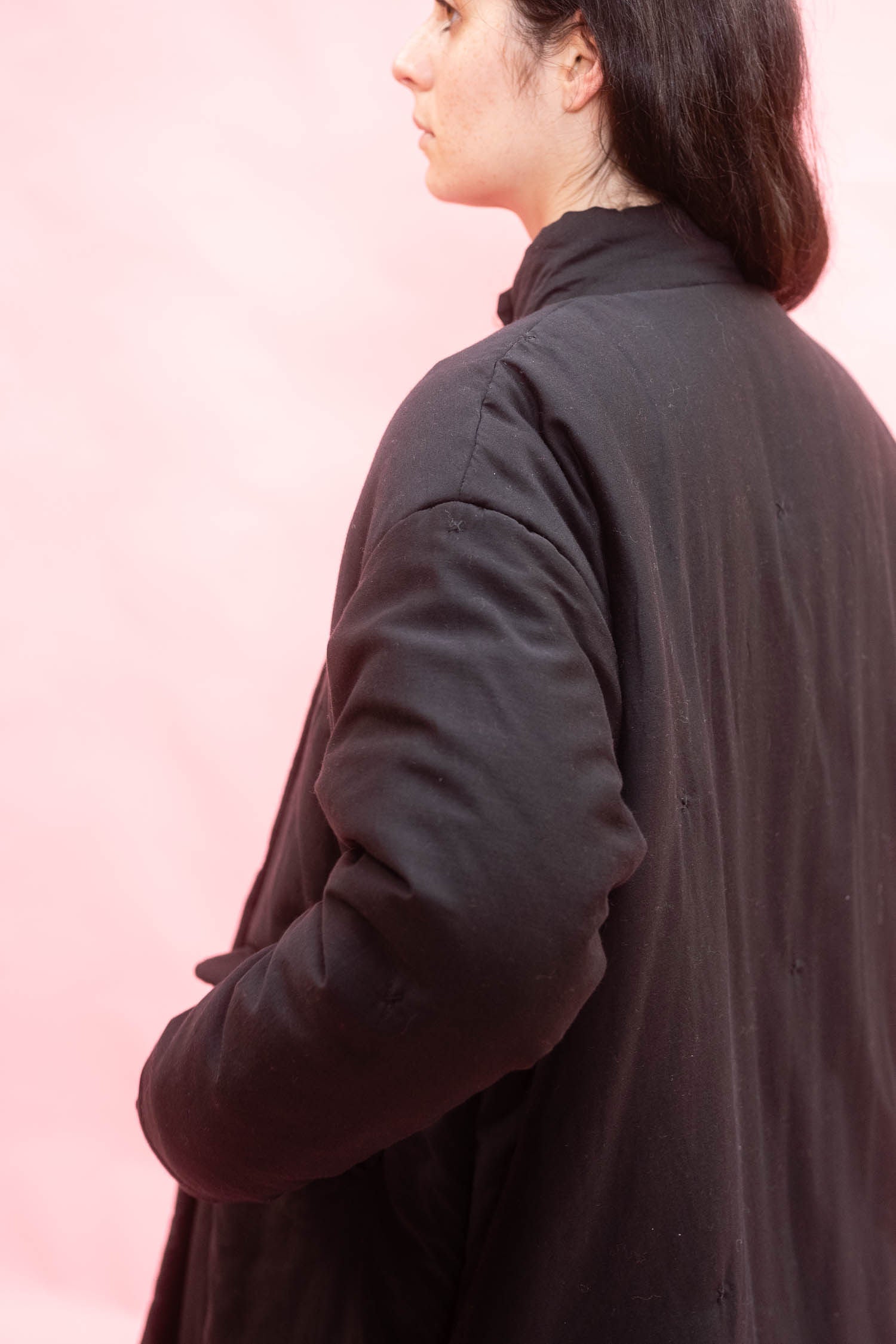 A woman shown from the back and side wearing an anthracite or black cuddle coat against a soft pink studio background.