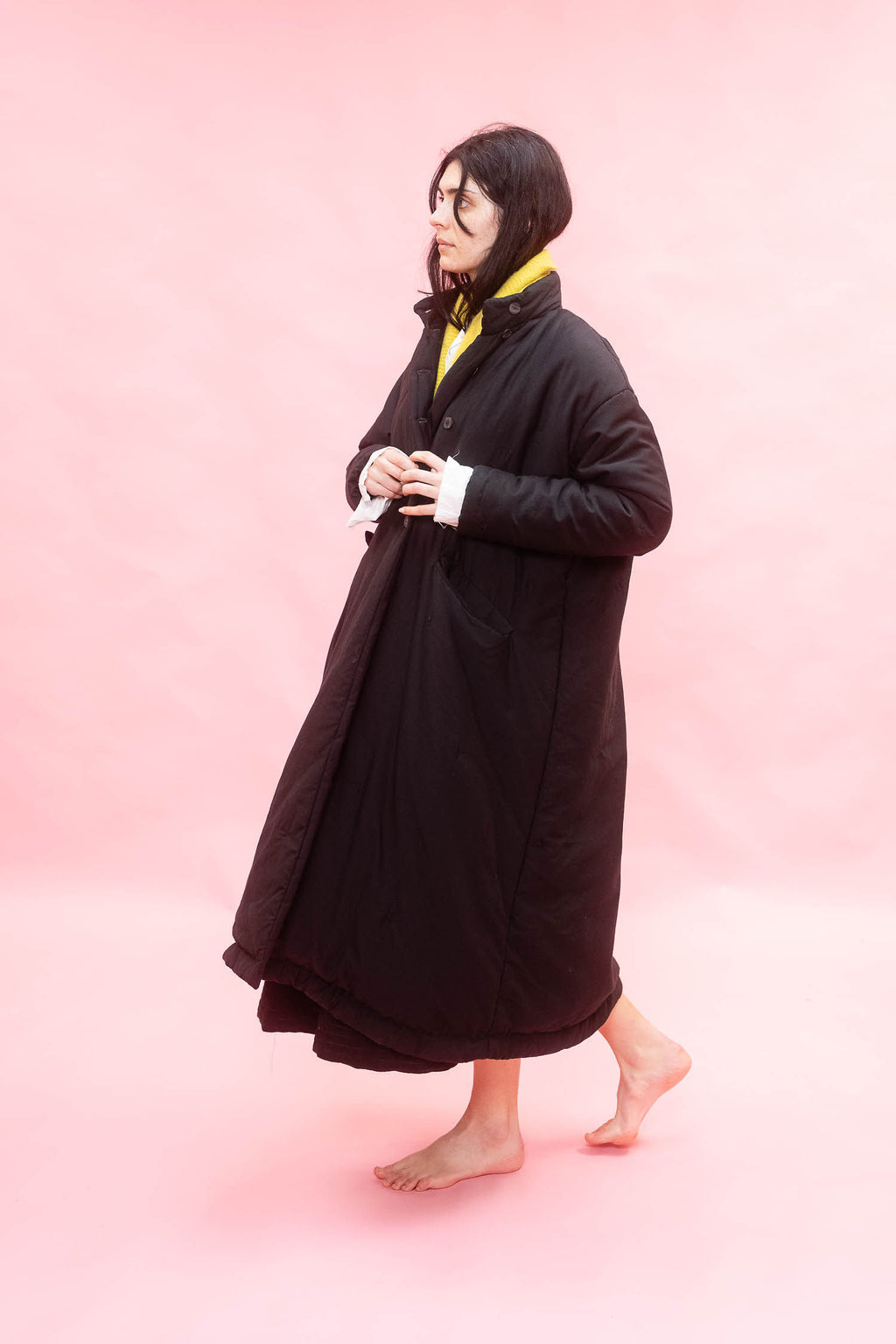 A woman wearing a long black quilted coat over a yellow scarf and white shirt, standing barefoot against a solid pink studio background.