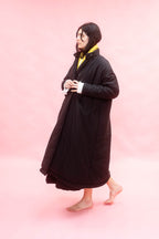 A woman wearing a long black quilted coat over a yellow scarf and white shirt, standing barefoot against a solid pink studio background.