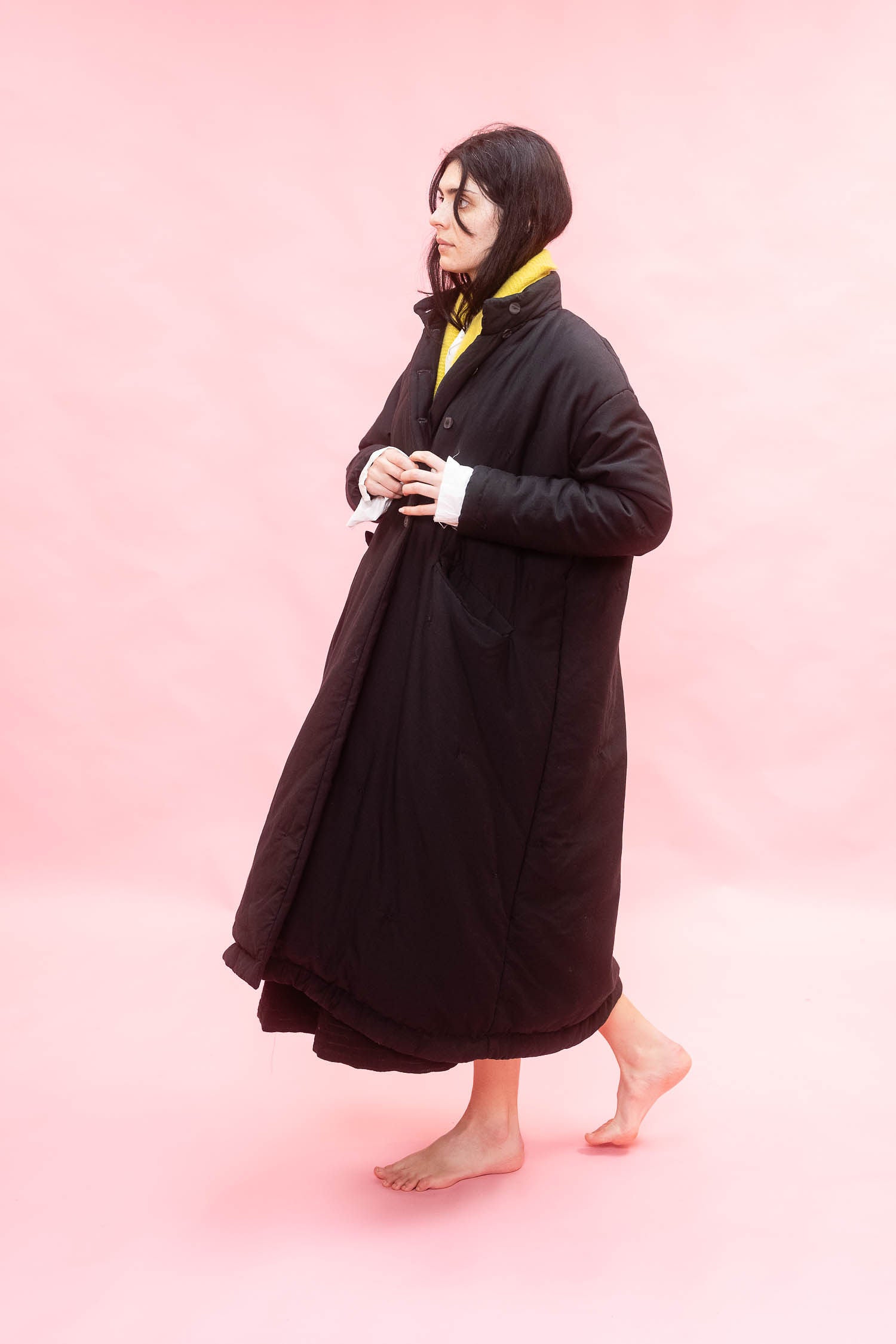 A woman wearing a long black quilted coat over a yellow scarf and white shirt, standing barefoot against a solid pink studio background.