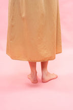 A person standing barefoot against a solid pink background, wearing a flowing, semi-sheer mustard yellow or ochre-colored dress or robe that drapes over the legs.