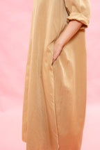 A close-up of a person wearing a flowing, lightweight tan dress or tunic with rolled sleeves, shown against a soft pink studio background.