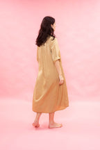 A woman with long dark hair is shown from behind wearing a loose, satin-like beige dress or robe with three-quarter sleeves, standing barefoot against a solid pink studio background.