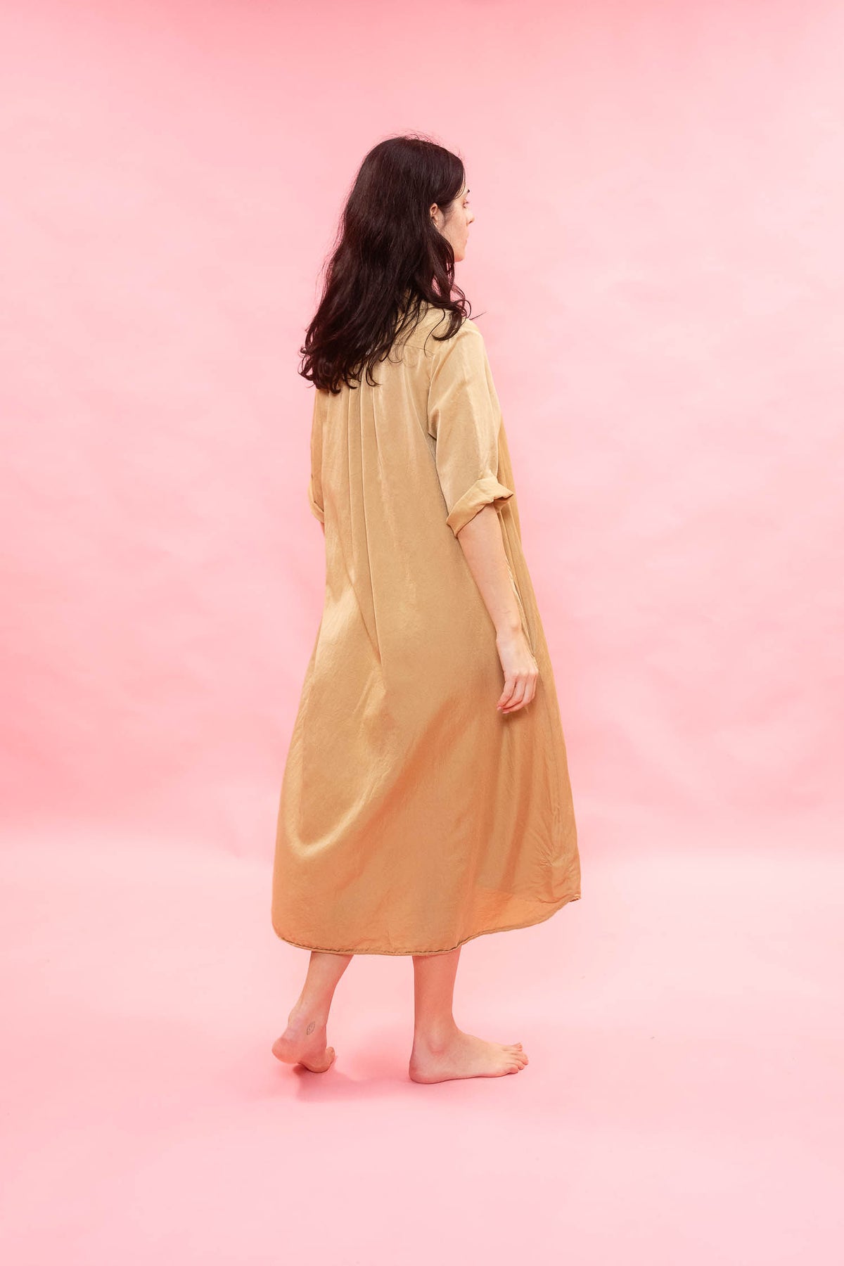 A woman with long dark hair is shown from behind wearing a loose, satin-like beige dress or robe with three-quarter sleeves, standing barefoot against a solid pink studio background.