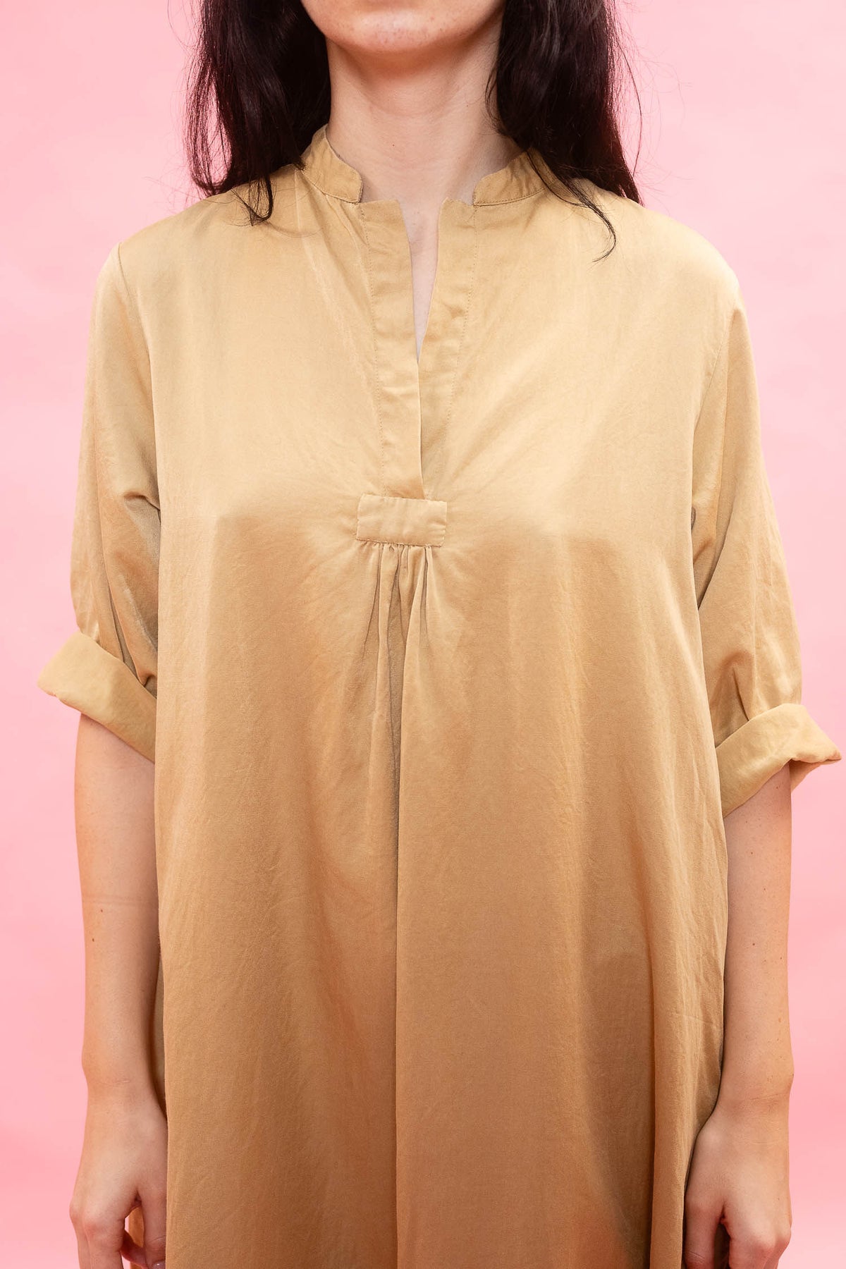 A woman wearing a loose, short-sleeved tan dress or tunic with a split neckline and gathered front detail, photographed against a solid pink background.