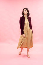 A woman stands barefoot against a solid pink studio background, wearing a dark burgundy cardigan over a light beige midi dress.