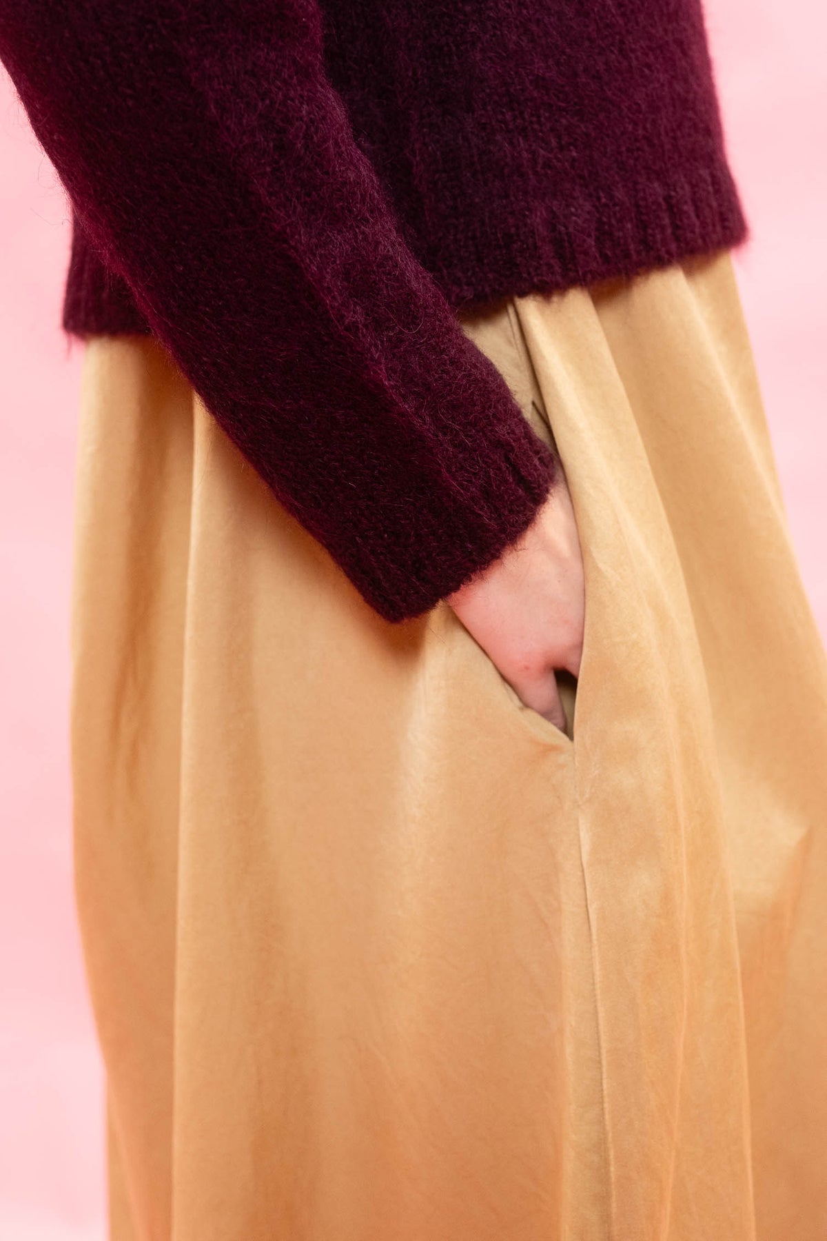 A close-up of a person wearing a dark burgundy fuzzy knit sweater and a flowing champagne-gold skirt, with one hand in the skirt pocket, set against a solid pink background.