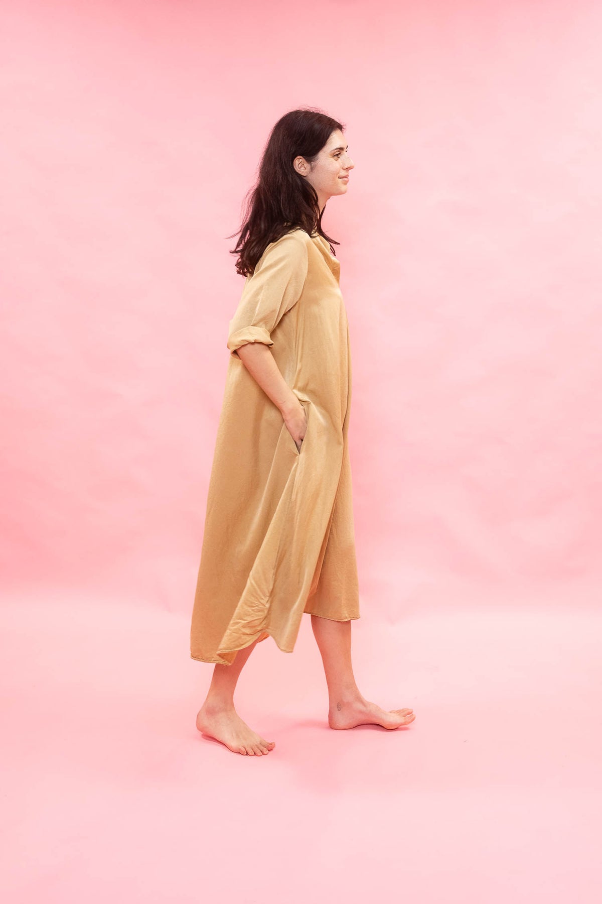 A woman stands in profile wearing a loose, knee-length beige linen dress against a solid pink studio background.