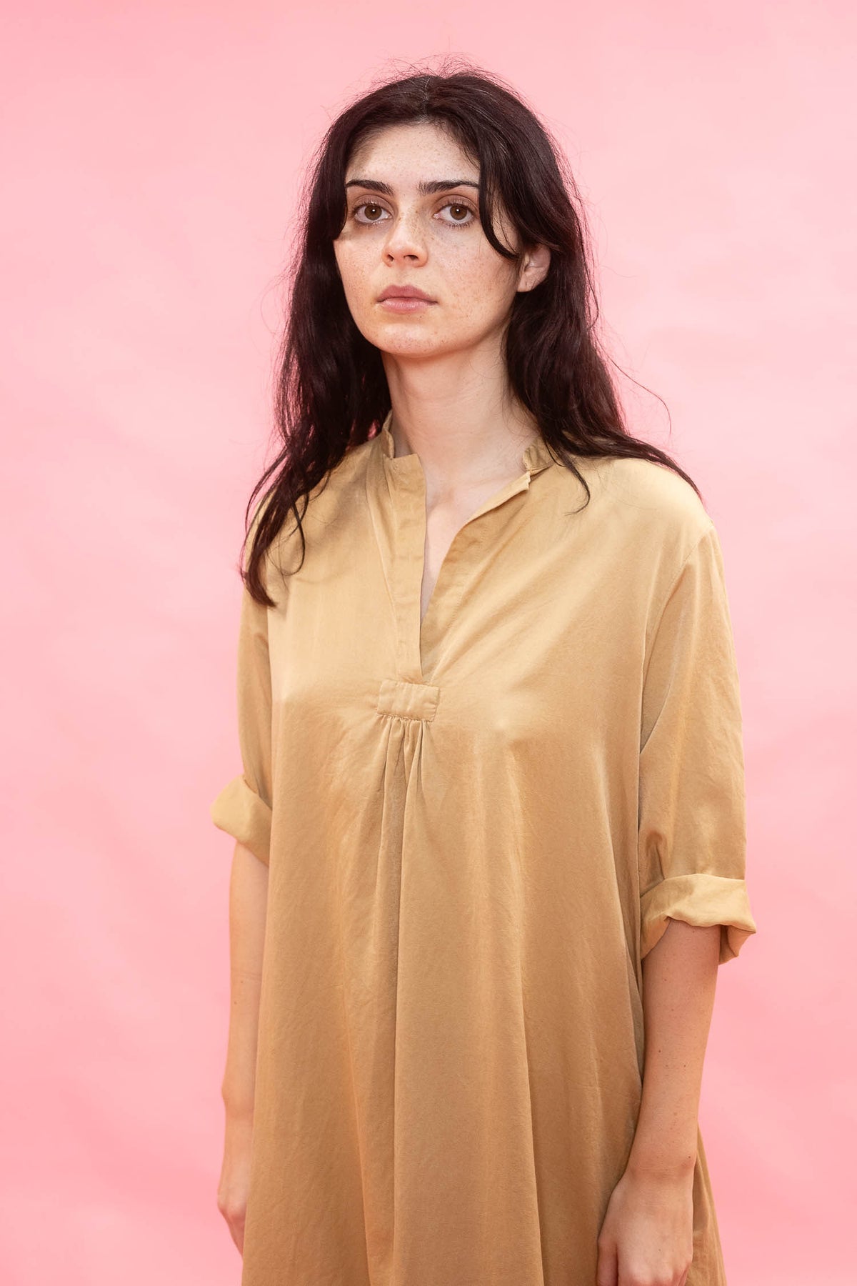A woman with long dark hair wearing a loose, short-sleeved mustard yellow tunic dress, standing against a solid pink background.