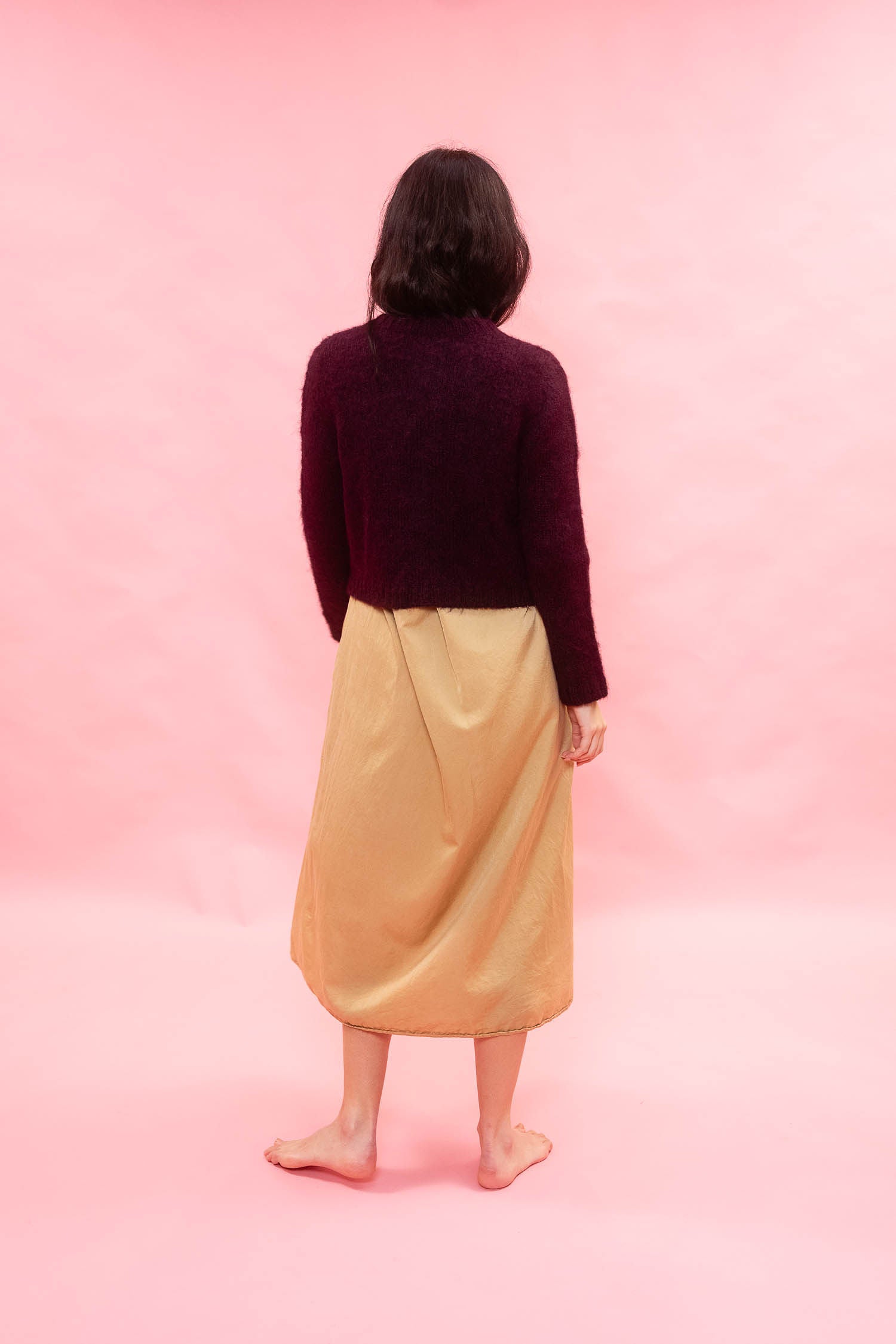 A person standing with their back to the camera, wearing a dark plumknit cropped alpaca cardiga nand a shiny mustard yellow dress, set against a solid pink studio background.

