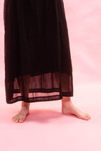 A person wearing a long black sheer maxi skirt with a sheer mesh hem, standing barefoot on a solid pink background.