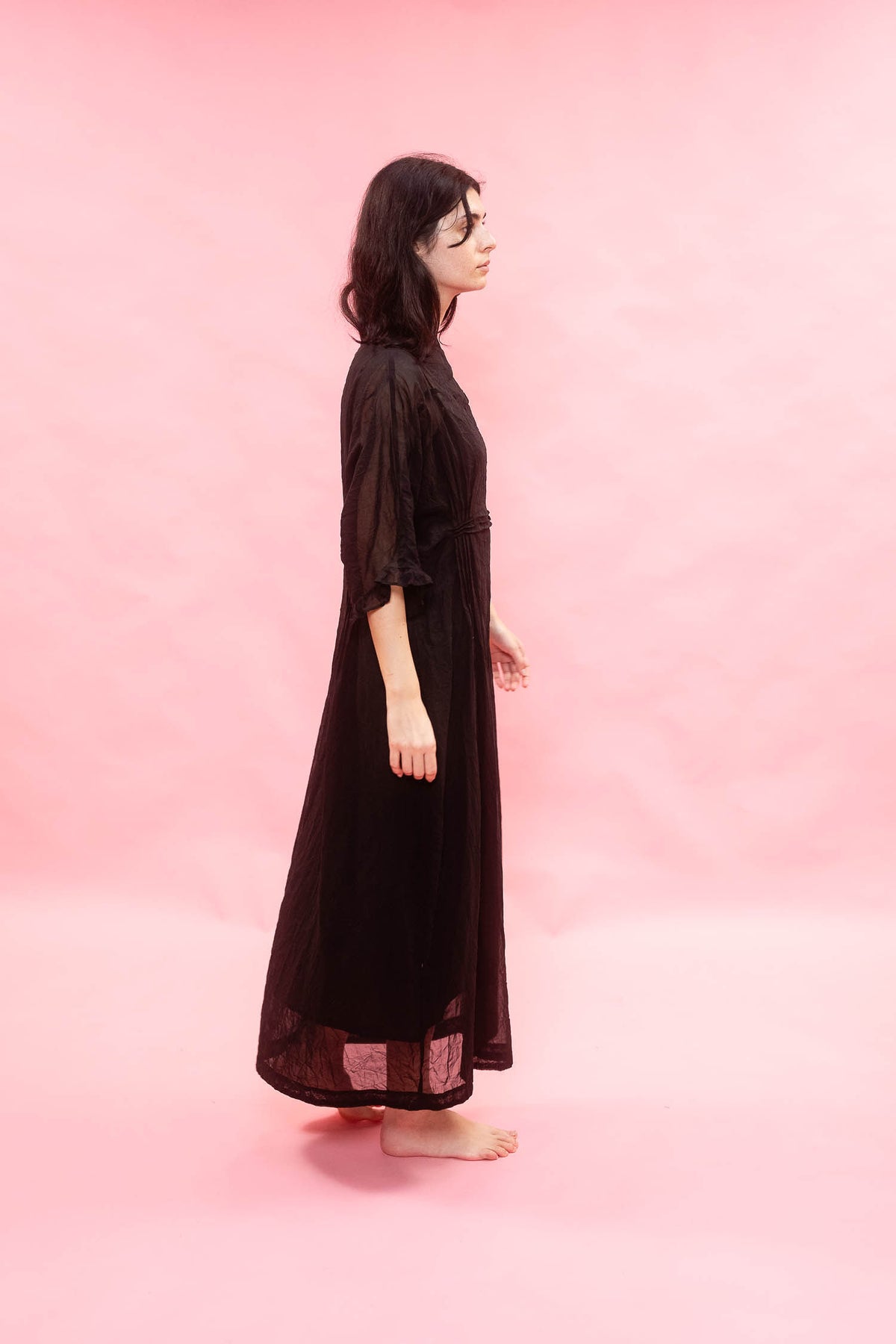 A woman stands in profile wearing a long black sheer maxi dress with elbow-length sleeves, barefoot against a solid pink studio backdrop.