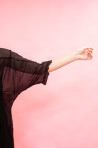 A person wearing a sheer black long-sleeve top or robe, shown from the side with one arm extended, against a solid pink background.
