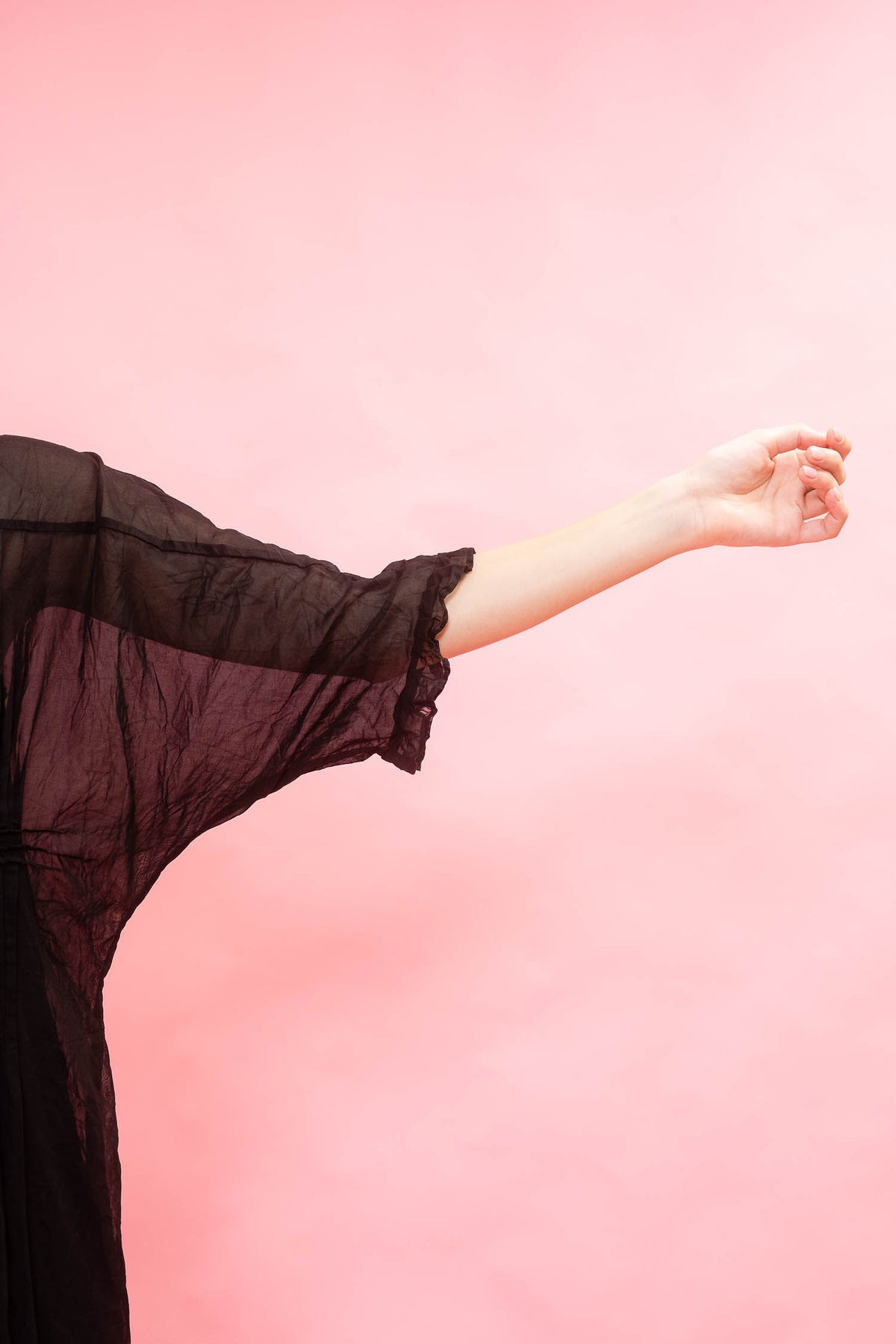 A person wearing a sheer black long-sleeve top or robe, shown from the side with one arm extended, against a solid pink background.