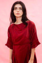 A woman with dark hair wearing a deep red, loose-fitting dress with three-quarter sleeves with a frill on the cuff, standing against a solid pink background.