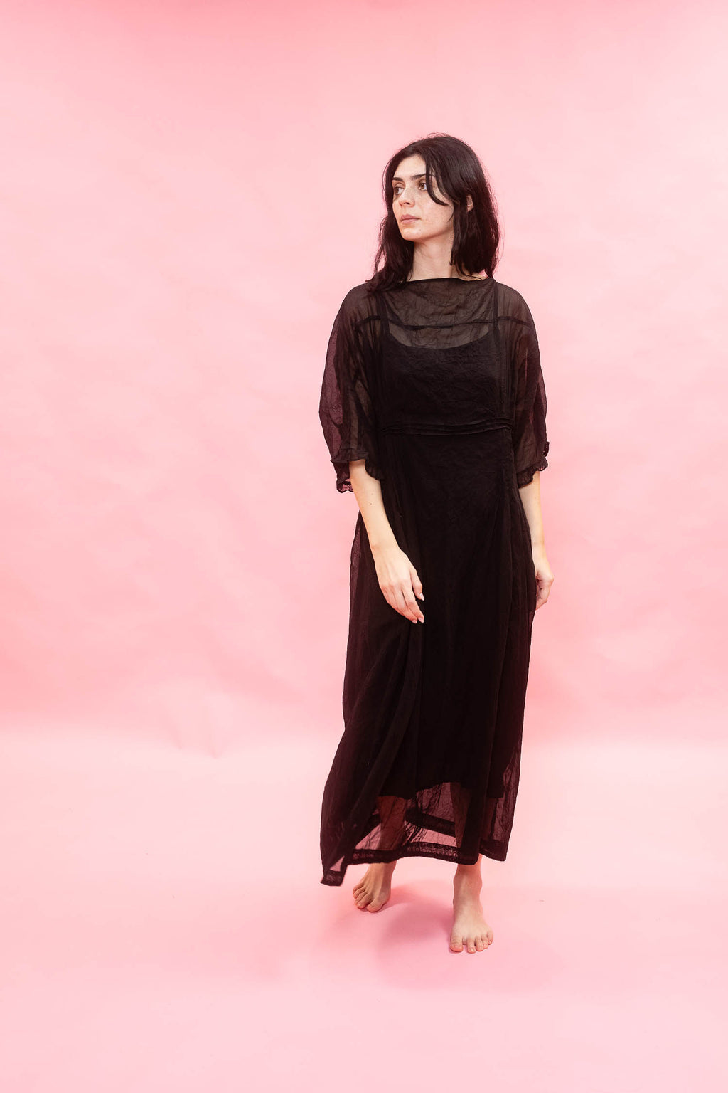 A woman wearing a long black sheer maxi dress with elbow-length sleeves, standing barefoot against a solid pink studio backdrop.