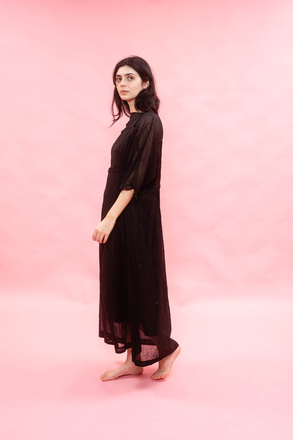 A woman stands barefoot in profile wearing a long, loose black sheer maxi dress against a solid pink studio backdrop.