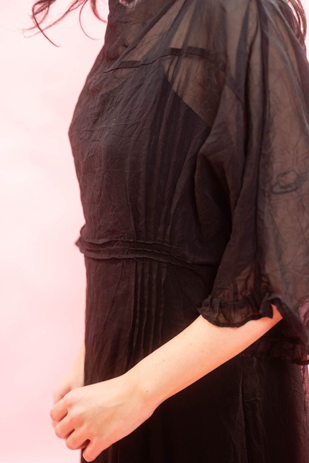 A close-up of a person wearing a black sheer mesh overlay dress with a gathered waist and ruffled elbow-length sleeves, set against a solid pink background.